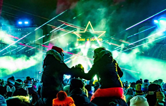 Deux personnes sur les épaules de deux autres personnes qui se tiennent par la main au festival Igloofest, devant la scène.