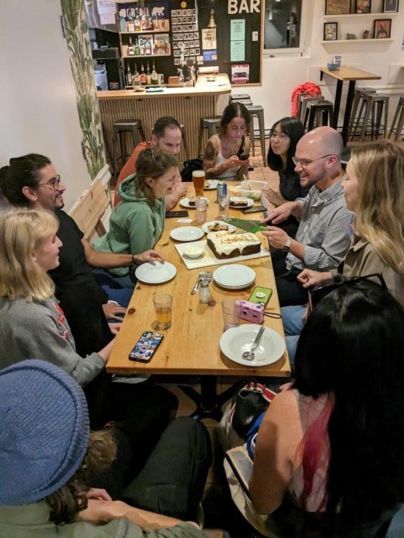 Plusieurs personnes autour d'une table qui mangent ensemble lors d'un souper communautaire à l'auberge Saintlo Montréal.