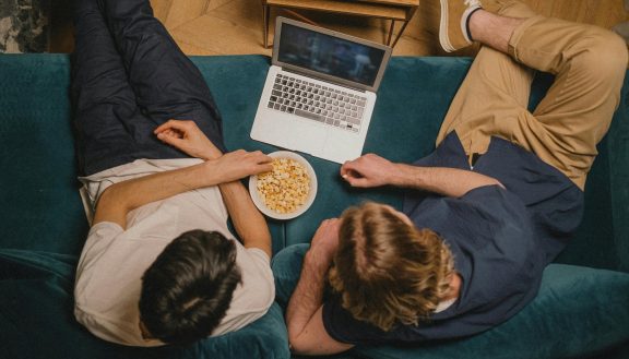 Deux personnes sur un sofa regardent un film sur un ordinateur portable en mangeant du popcorn.